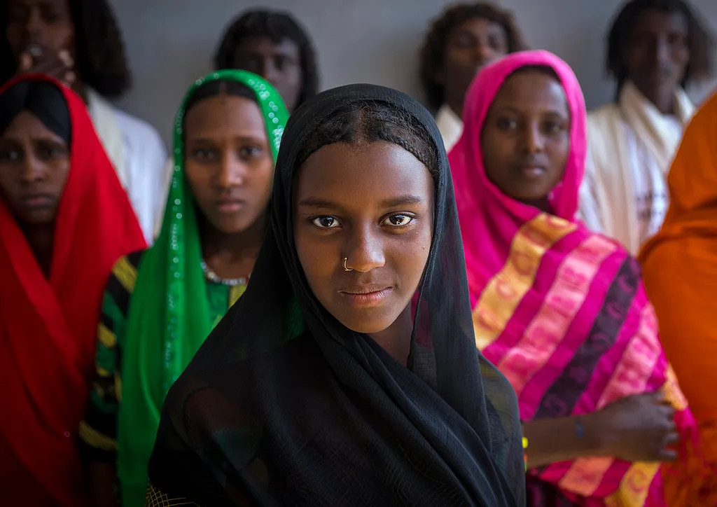 A young Afar woman in a black headscarf stands in front of a group of women in brightly coloured scarves